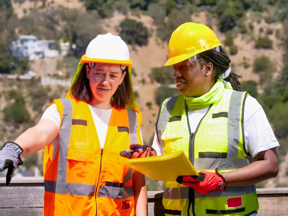 Two safety professionals in hard hats and high-visibility vests reviewing plans at a construction site